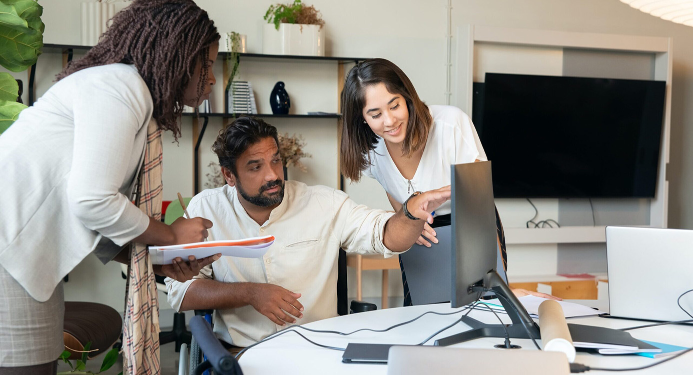 Three people of colour are looking at a computer screen in a workplace. The person in the middle is pointing at the screen and in a manual wheelchair.